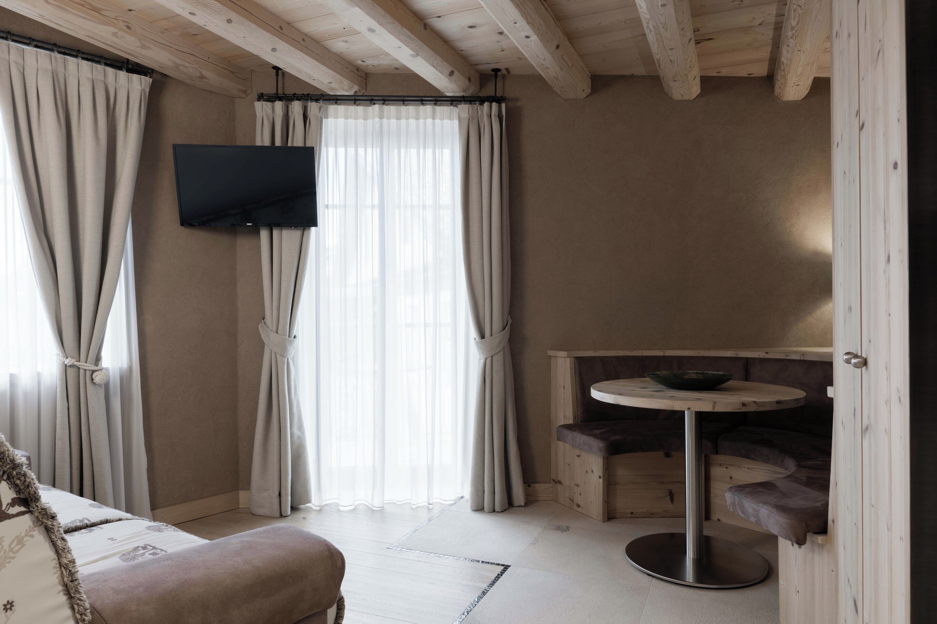 A room with a television mounted on the wall, a round wooden table, a cushioned bench, and a window with curtains.