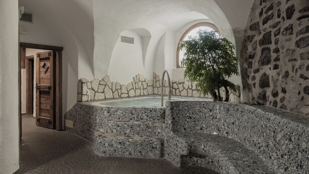Zona de piscina con paredes de piedra, escalones que conducen a un área de descanso, una puerta de madera y un árbol visible a través de una ventana.