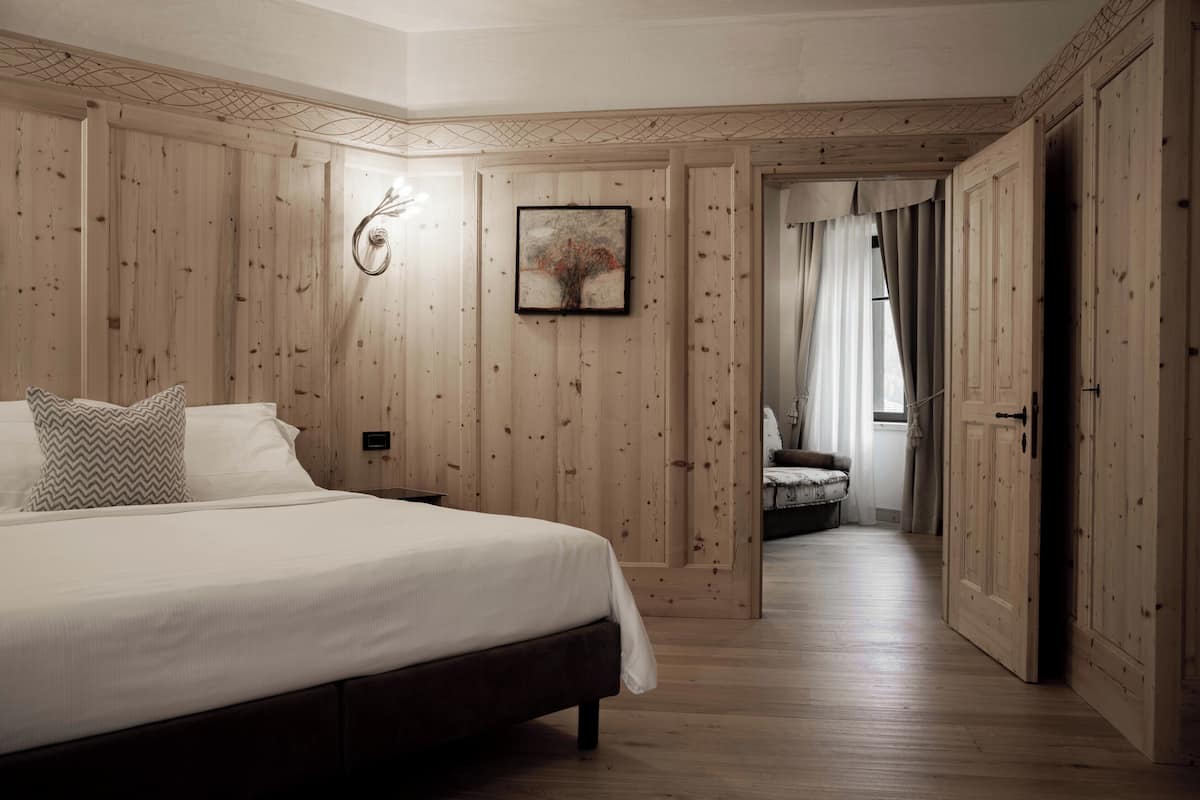 A bedroom with a wooden paneled wall, a bed with white bedding, a framed artwork, and a door leading to another room.