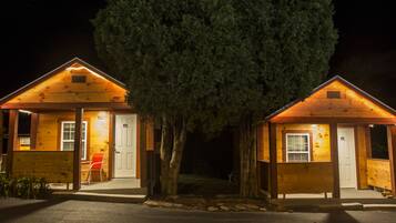 Two wooden cabins with a large tree in front, each with a door and window, and a chair outside.