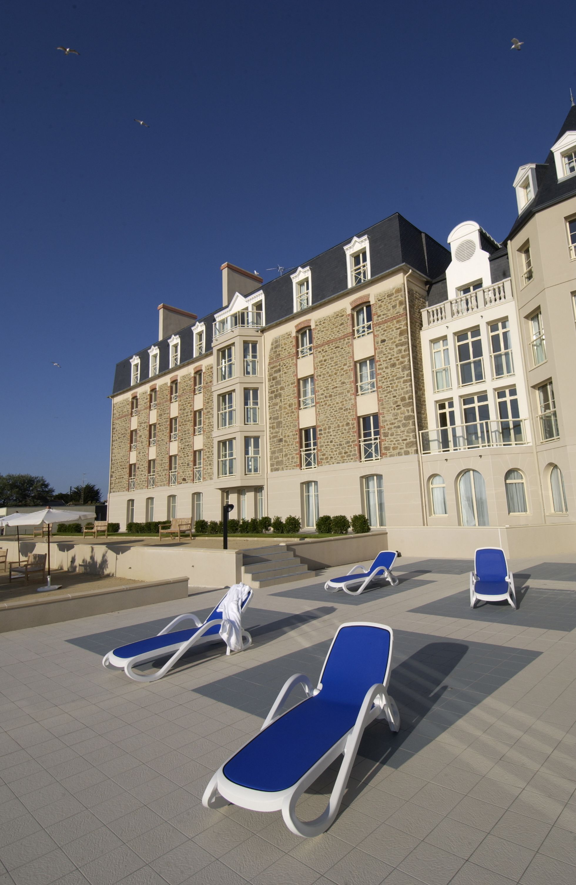 A hotel with a stone facade, multiple balconies, and a pool area with lounge chairs.