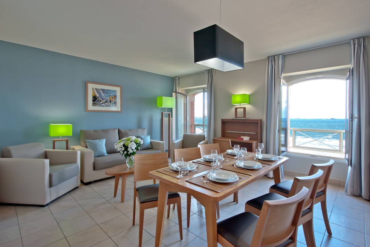 A modern hotel room with a dining area, a sofa set, and a large window offering a view of the sea.