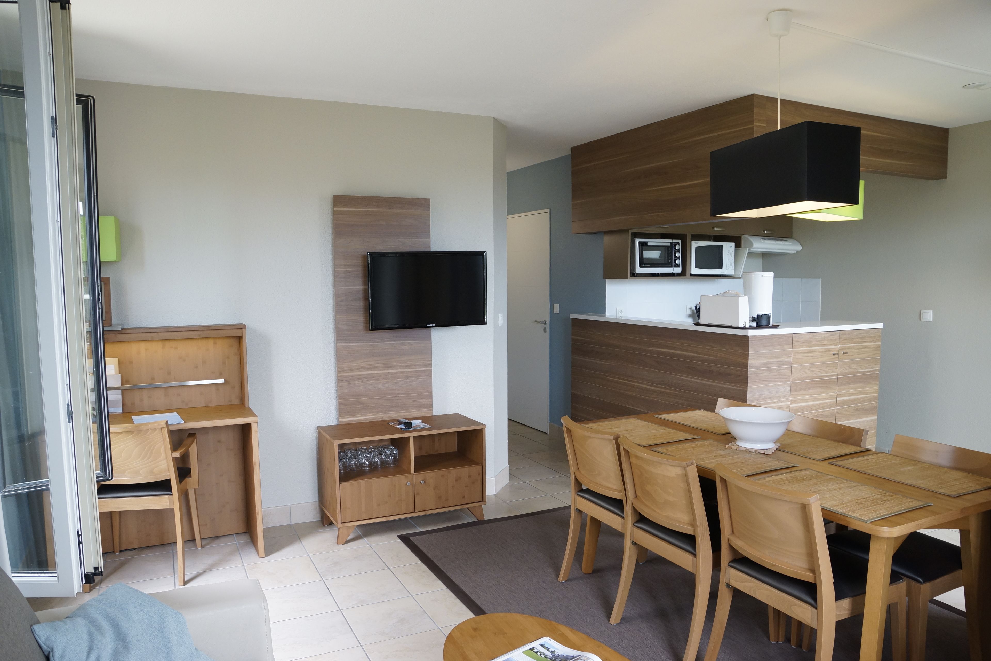 Ein modernes, gut beleuchtetes Zimmer mit Schreibtisch aus Holz, Fernseher, Esstisch mit Stühlen und einer Küchenzeile mit Mikrowelle und Kaffeemaschine.