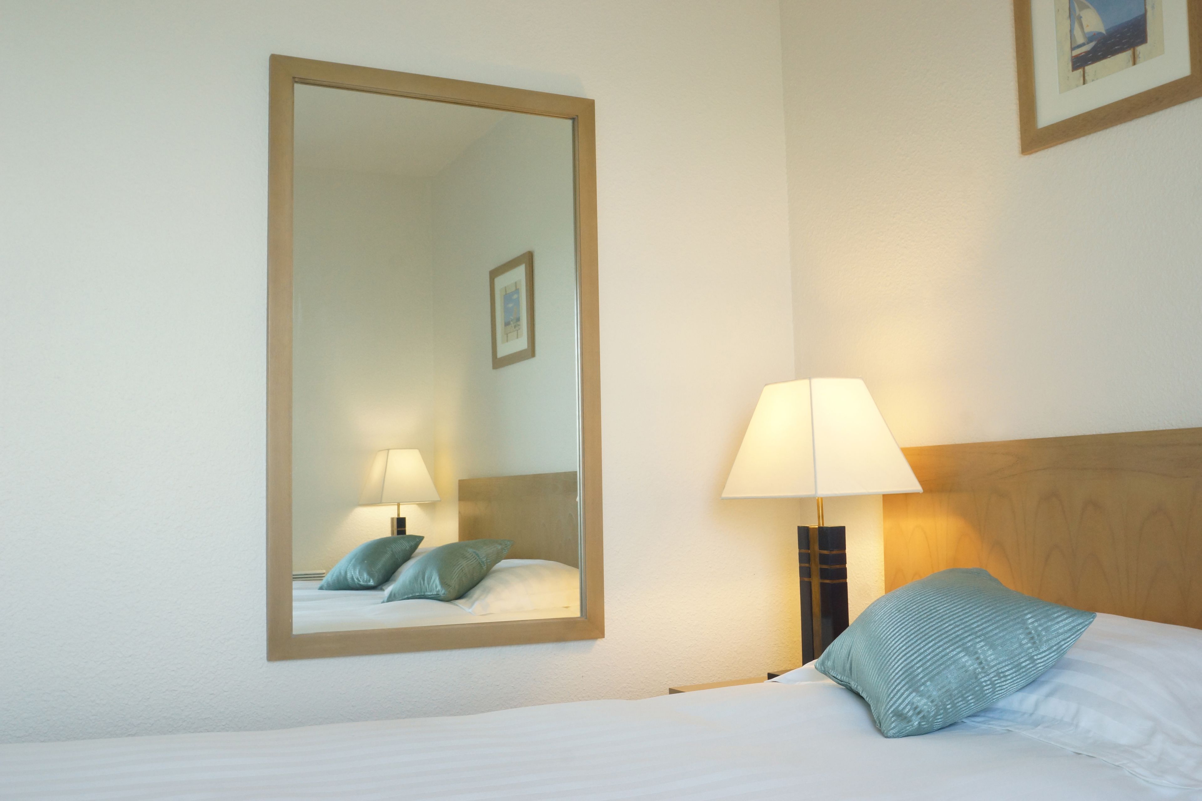 A hotel room with a large mirror, a bed with a pillow, a nightstand with a lamp, and a framed picture on the wall.