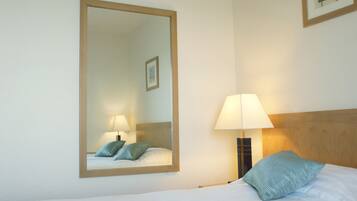 A hotel room with a large mirror, a bed with a pillow, a nightstand with a lamp, and a framed picture on the wall.