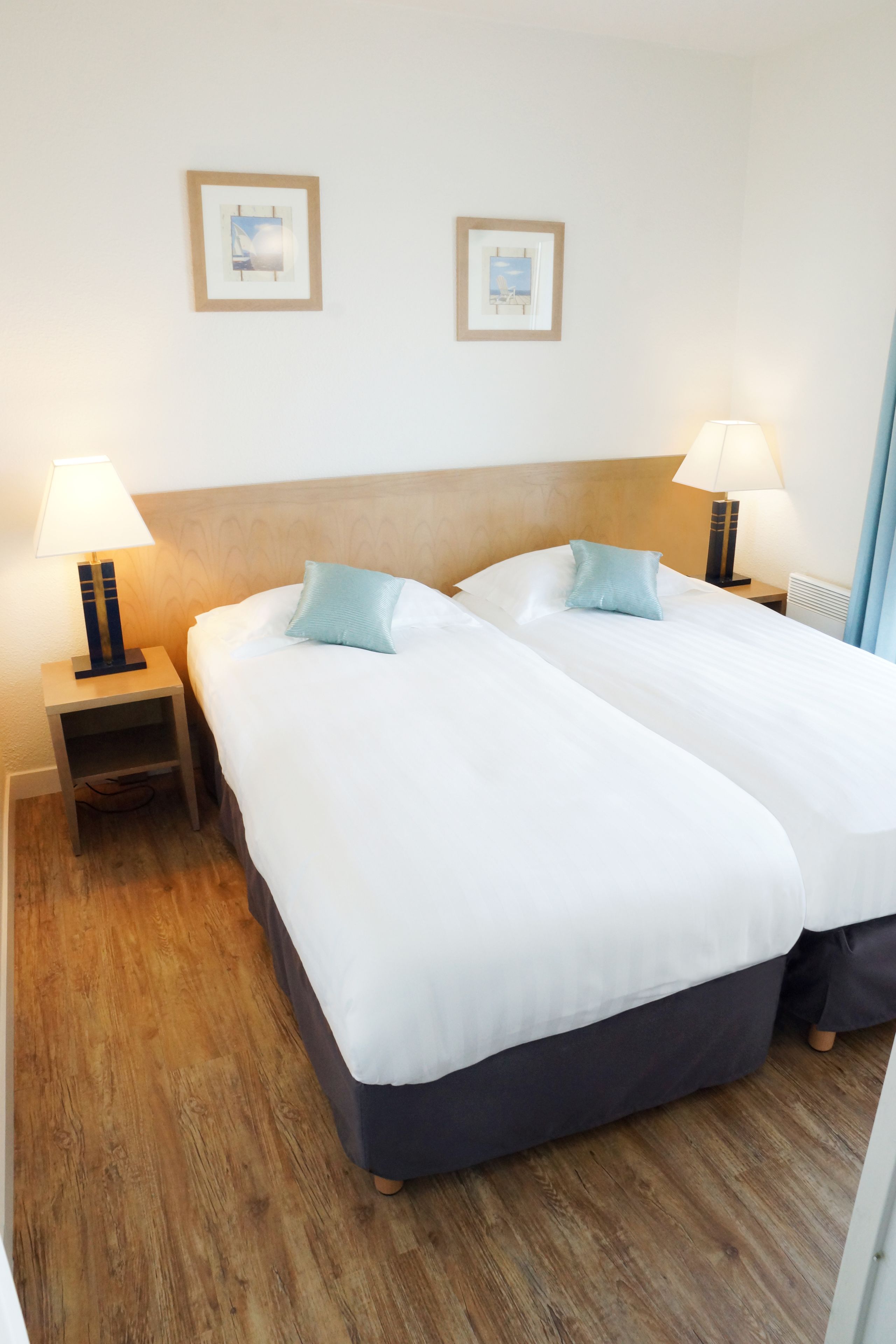 A hotel room with two beds, wooden headboard, bedside tables with lamps, and framed pictures on the wall.