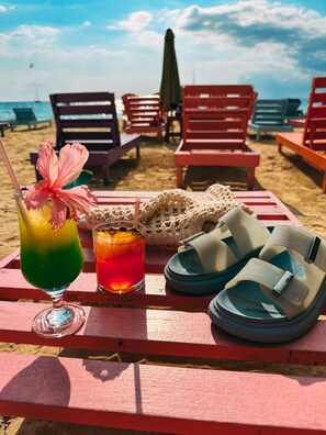 Une scène de plage avec une table sur laquelle on aperçoit une boisson colorée, un article en crochet et une paire de sandales.