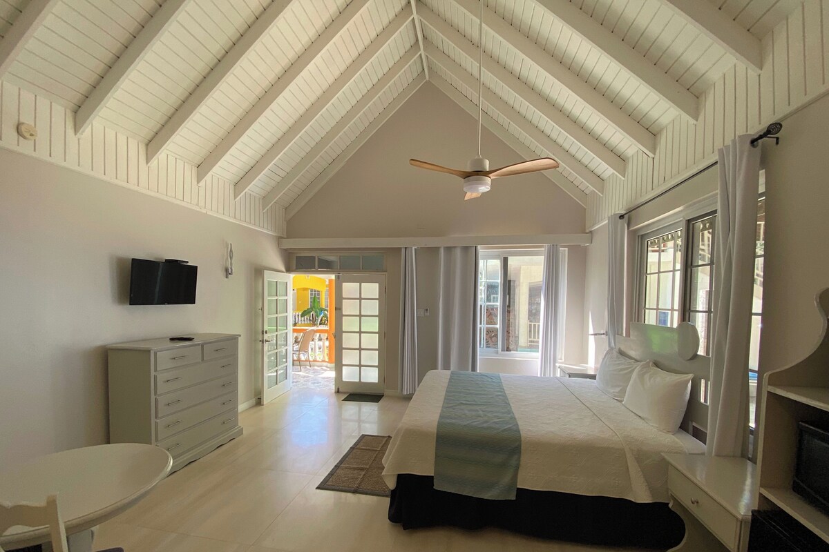 A spacious bedroom with a high ceiling, a bed with a headboard, a bedside table, a dresser, a television mounted on the wall, and a window with curtains.