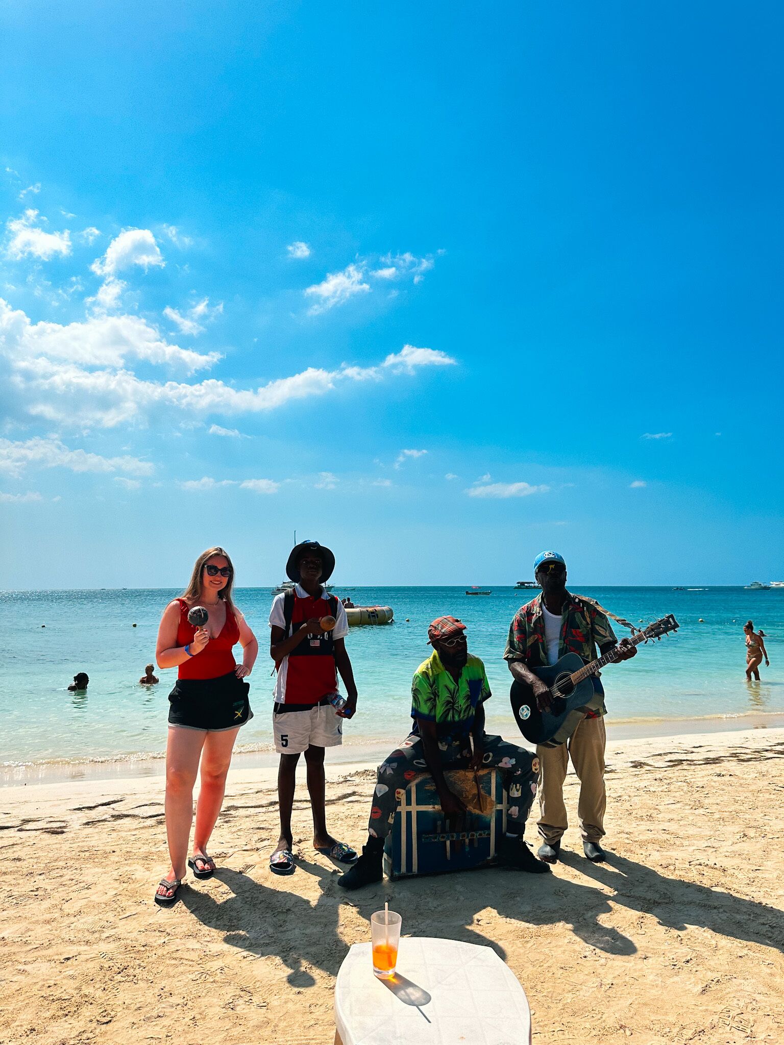 Escena de playa con gente, un guitarrista y una mesa con una bebida.