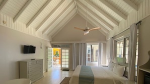 A spacious bedroom with a high ceiling, a bed with a headboard, a bedside table, a dresser, a television mounted on the wall, and a window with curtains. - The Boardwalk Village (Negril)