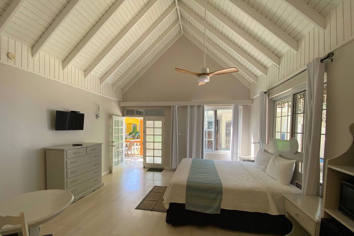 A spacious bedroom with a high ceiling, a bed with a headboard, a bedside table, a dresser, a television mounted on the wall, and a window with curtains.