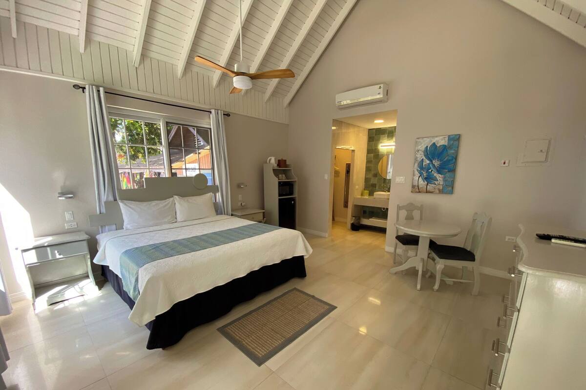 A hotel room with a bed, a desk, a chair, a dining table, and an air conditioning unit.