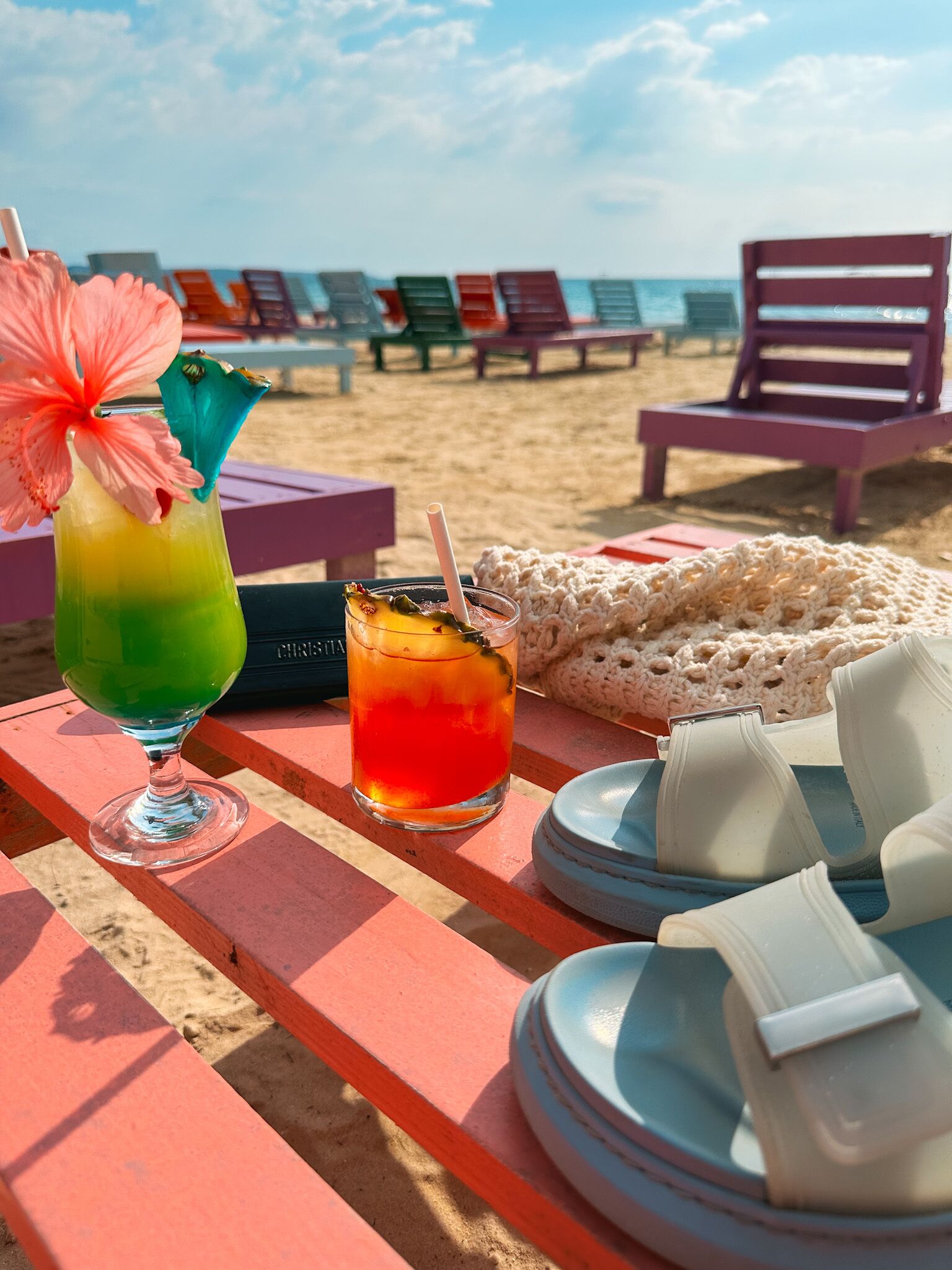 Una mesa en la playa con dos bebidas, una manta de crochet y sillas de playa.