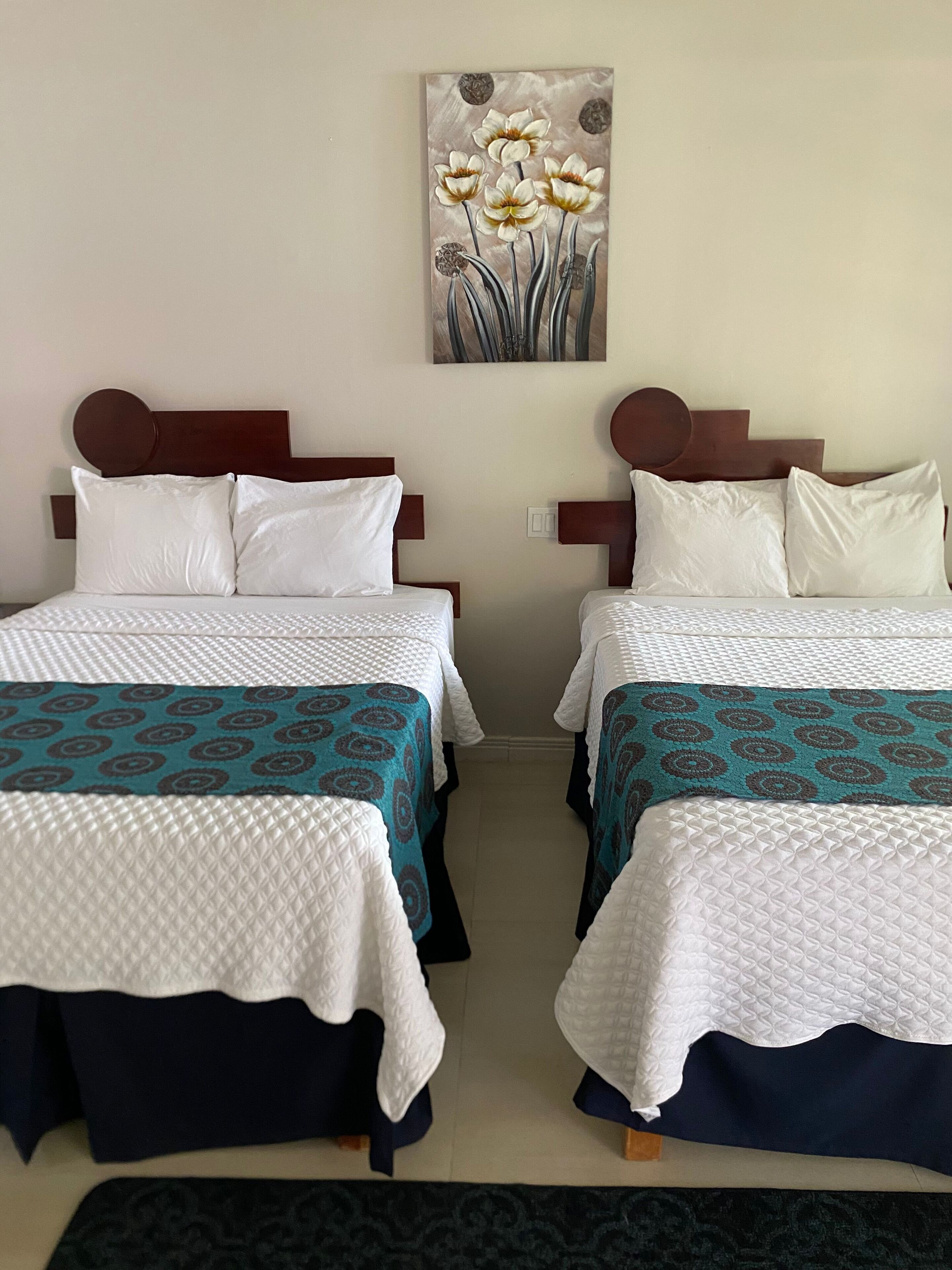 A hotel room with two beds, each with white bedding and teal patterned bedspreads. There is a painting of flowers on the wall above the beds.