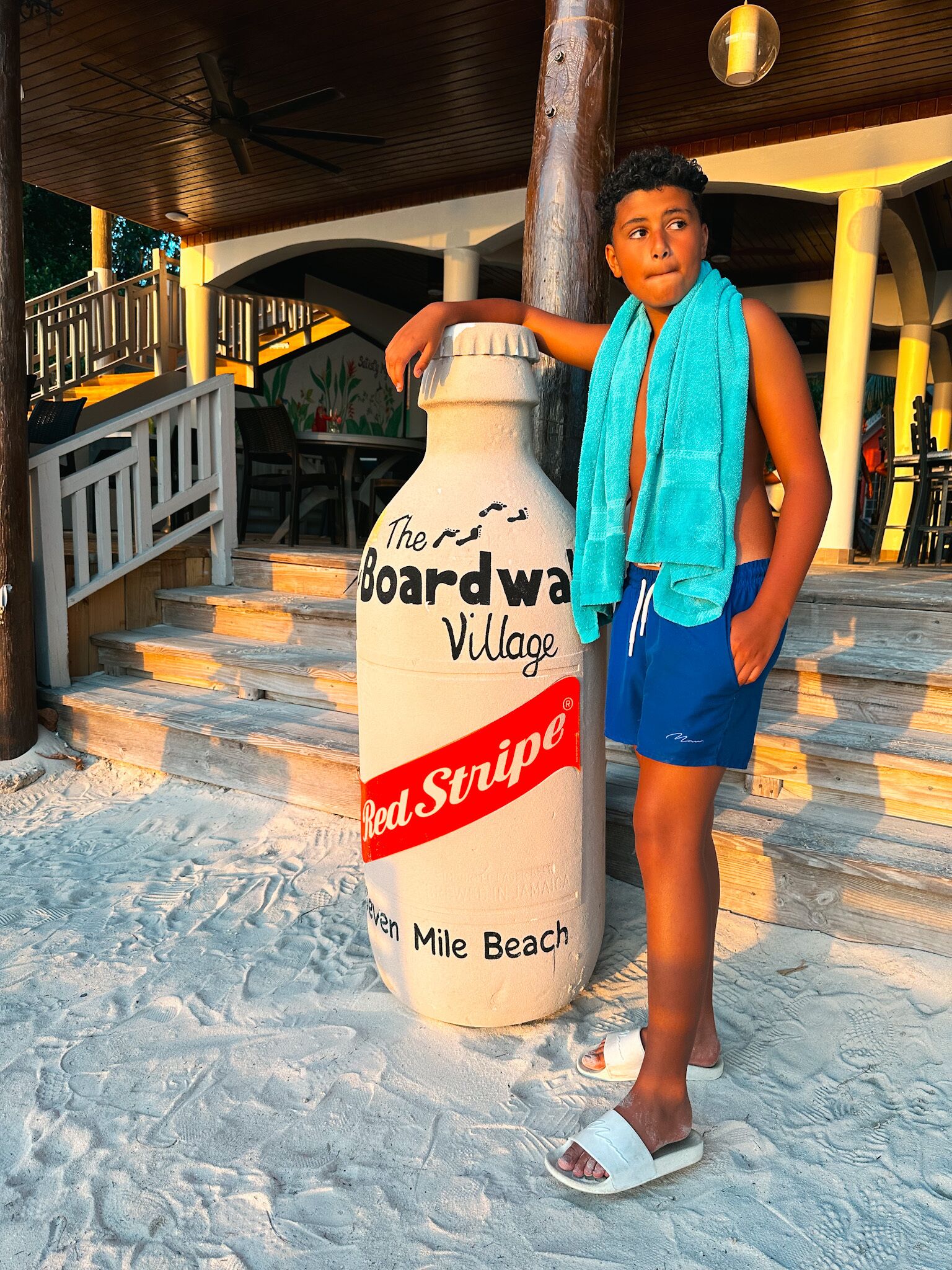 Eine Person steht neben einer großen Skulptur in Form einer Red Stripe Bierflasche auf sandigem Untergrund.