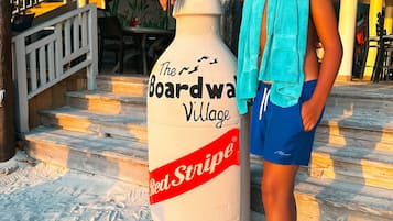 Eine Person steht neben einer großen Skulptur in Form einer Red Stripe Bierflasche auf sandigem Untergrund.