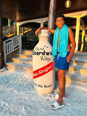 Une personne se tient à côté d’une grande sculpture en forme de bouteille de bière Red Stripe, posée sur une surface sablonneuse.