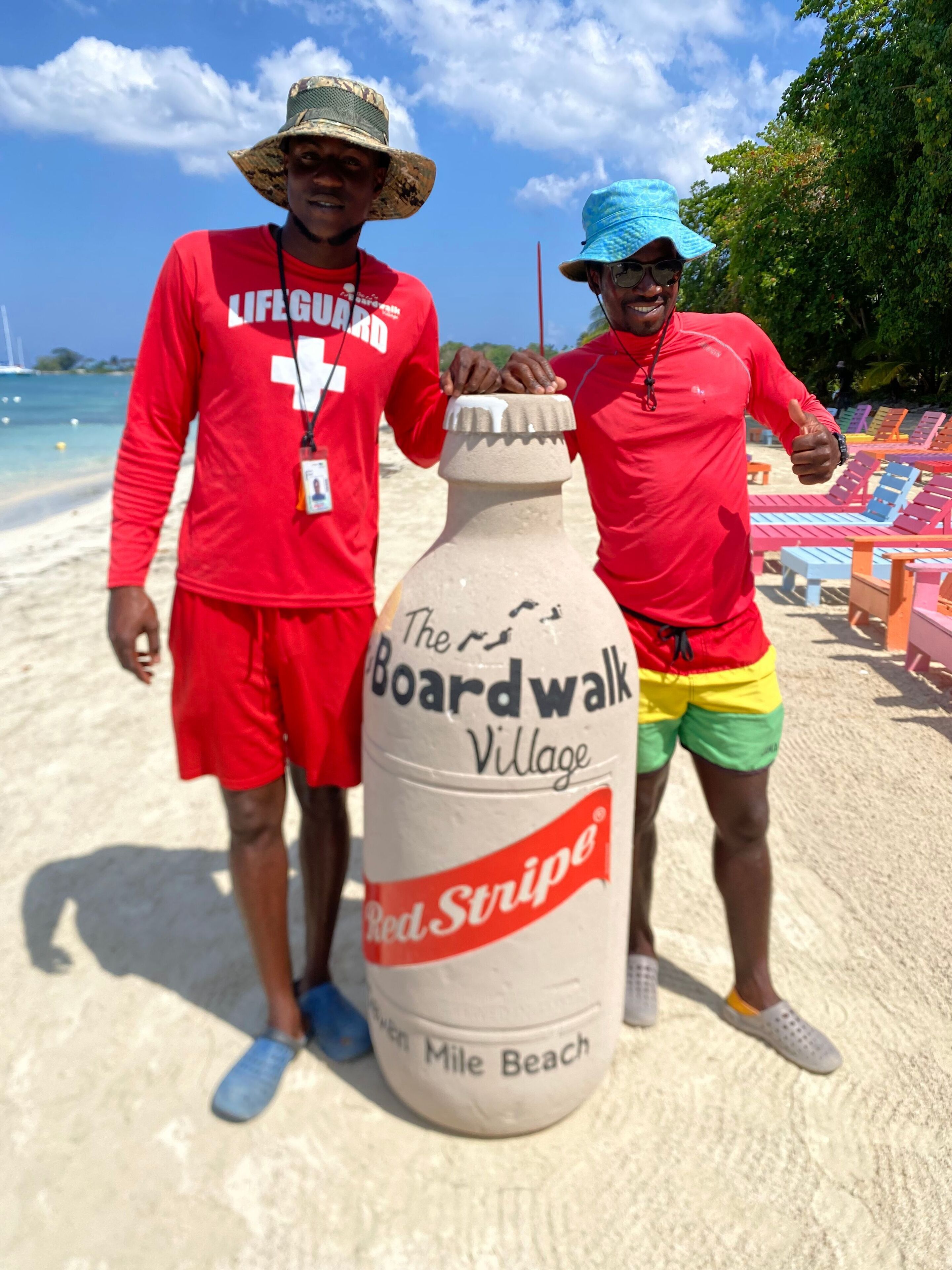 Two people standing on a sandy beach with a large bottle labeled "The Boardwalk Village Red Stripe" in the foreground.