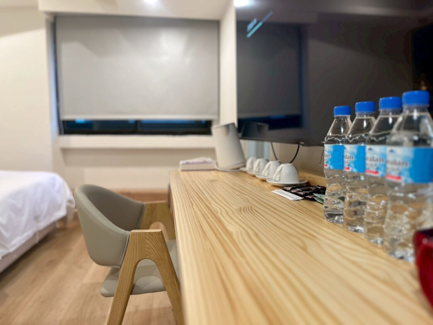 A hotel room with a wooden desk, a chair, a bed, a TV, and several water bottles.