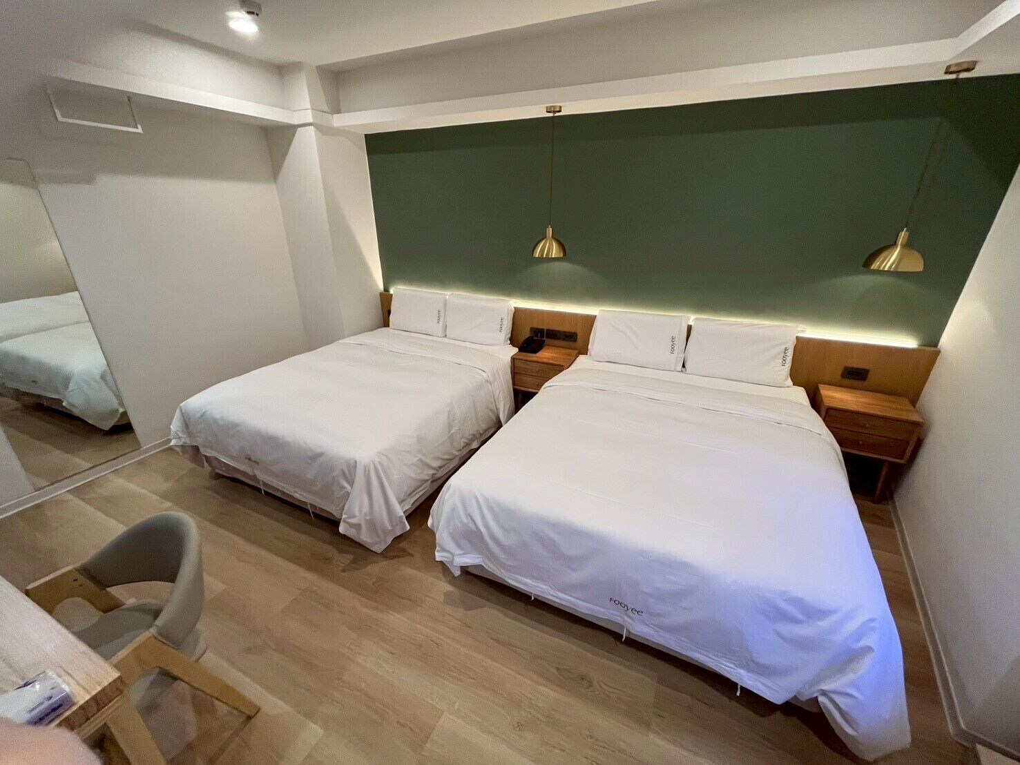 A hotel room with two single beds, wooden headboards, bedside tables, and a small desk with a chair.