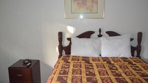 A bed with a patterned bedspread, a wooden headboard, and a nightstand with a clock. - Searle Motel (Long Beach)