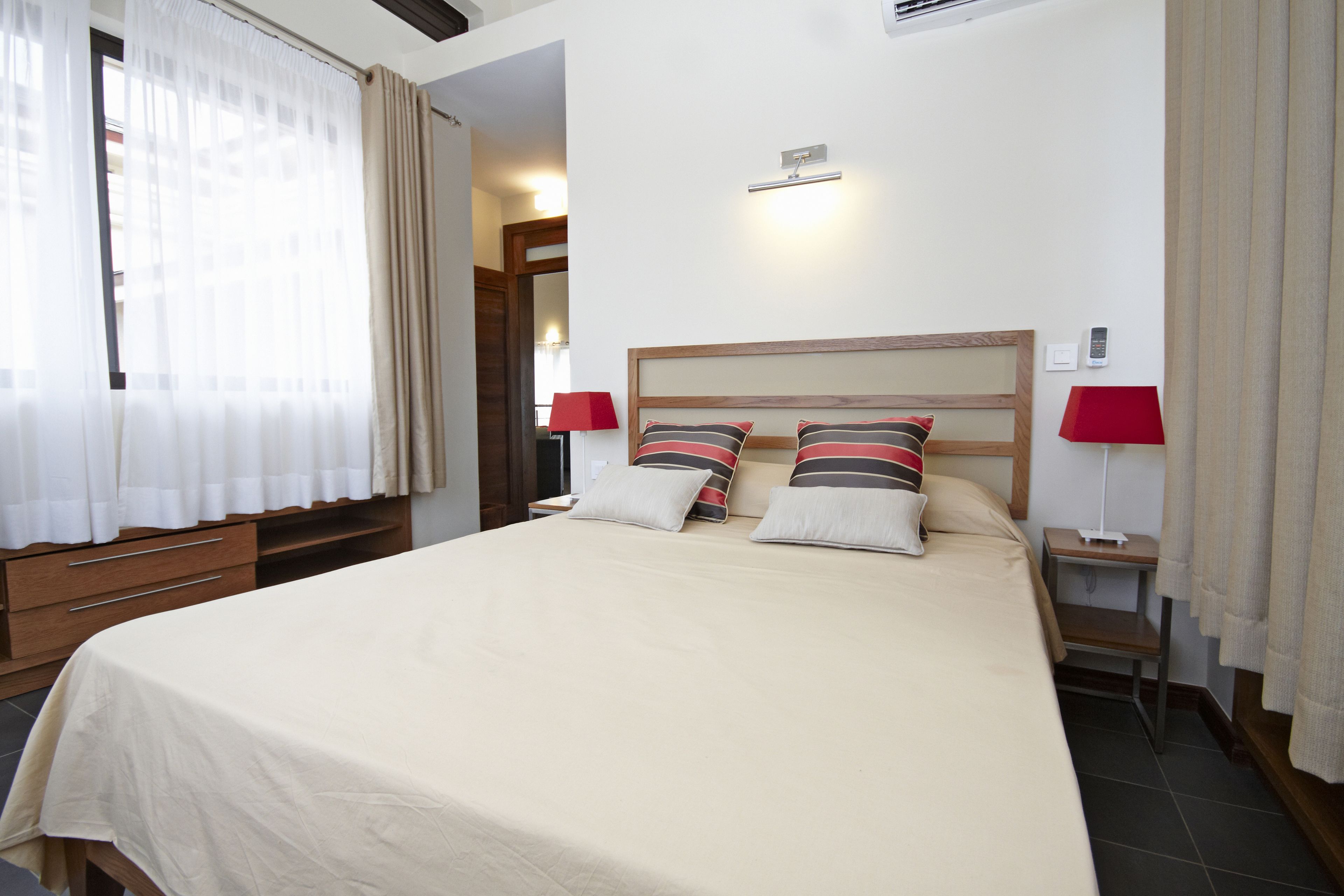 A hotel room with a large bed, two bedside tables, a nightstand, a wooden dresser, and a window with sheer curtains.