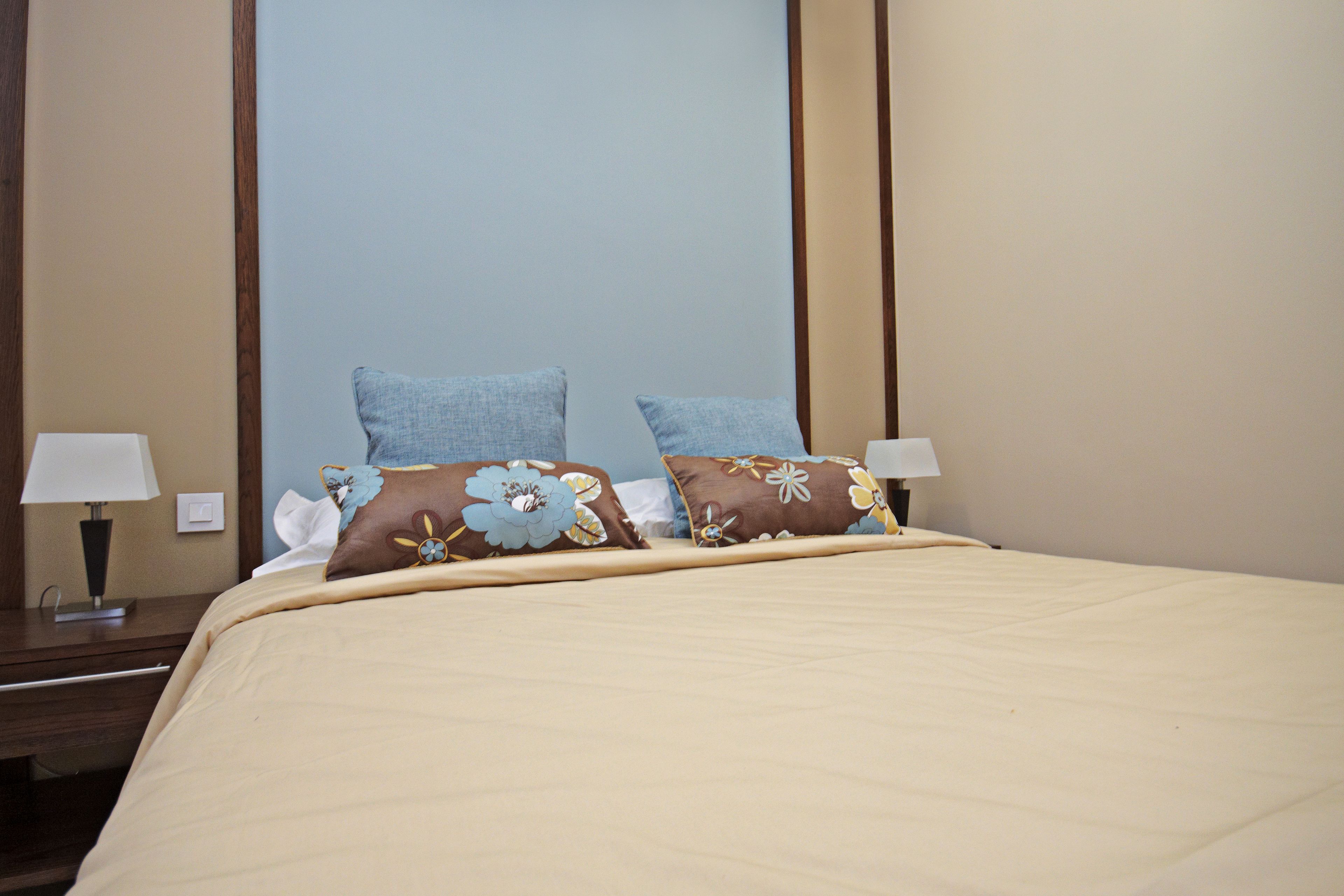 A neatly made bed with a beige bedspread and two decorative pillows, flanked by two bedside tables with lamps.