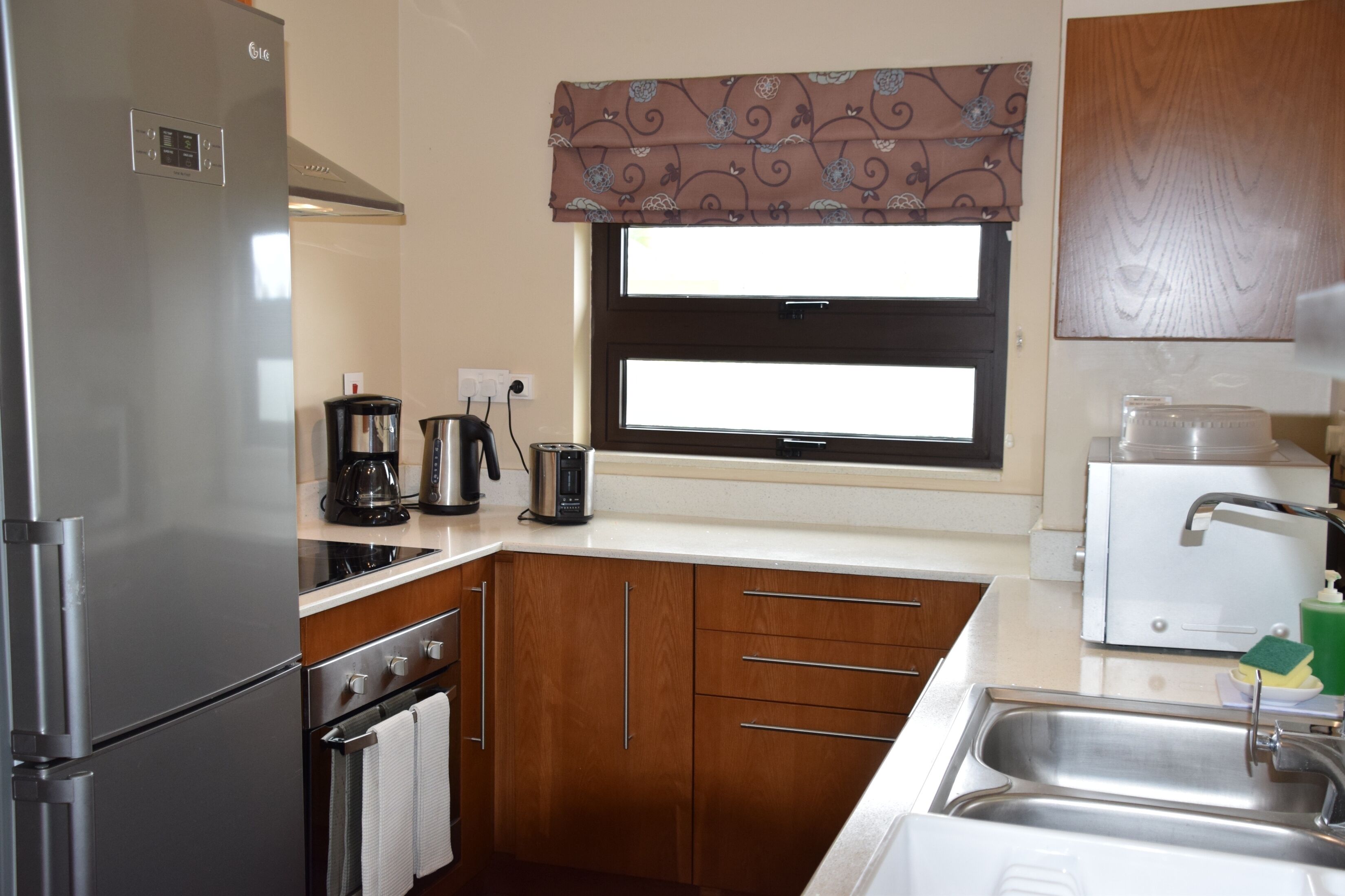 A compact kitchen with a refrigerator, stove, oven, and sink. There is a window with a patterned valance, and a microwave on the countertop.