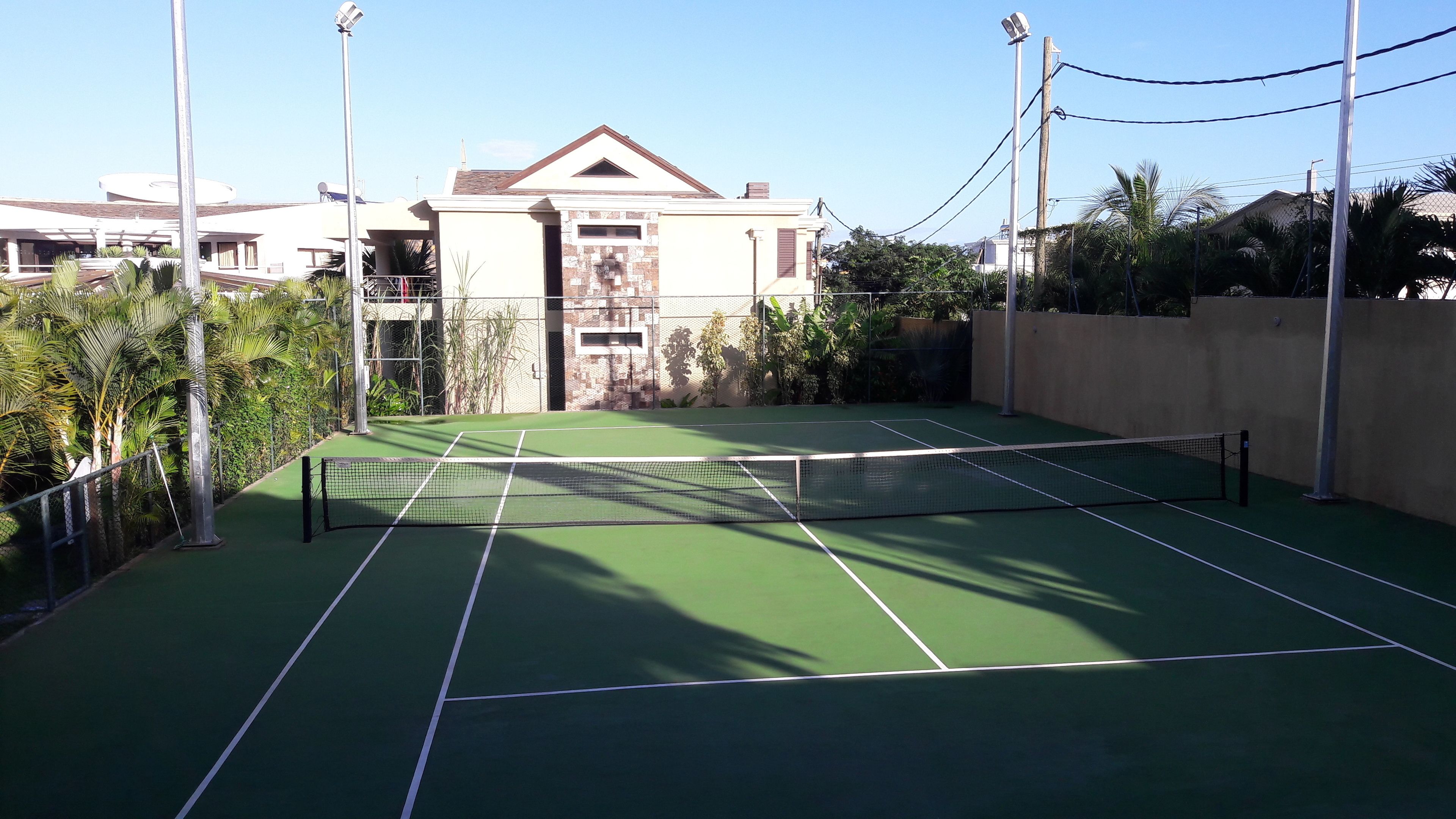 An outdoor tennis court with a net, surrounded by greenery and a building in the background.