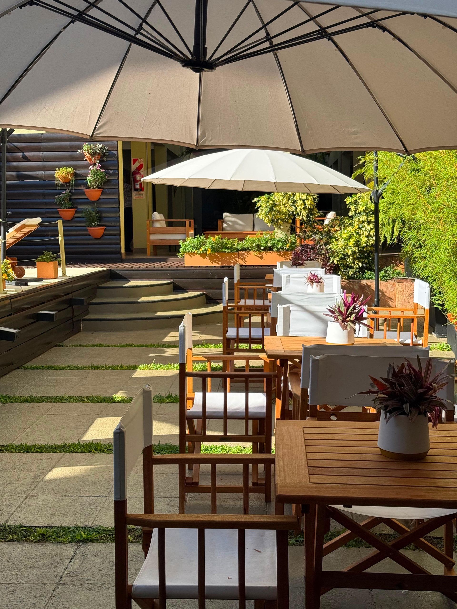 Un patio exterior con mesas y sillas de madera, un paraguas grande y plantas en macetas.