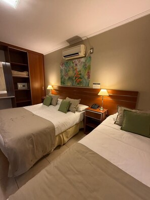 Habitación de hotel con dos camas, una mesita de noche con lámpara, aire acondicionado en la pared y un cuadro.