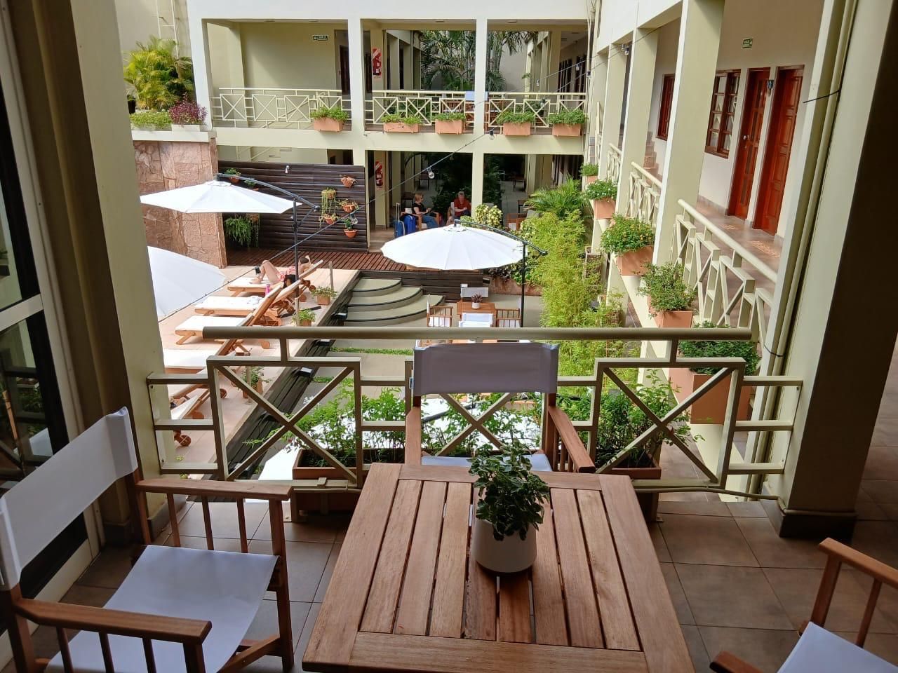 A balcony with wooden furniture, a potted plant, and a view of a courtyard with multiple levels and balconies.