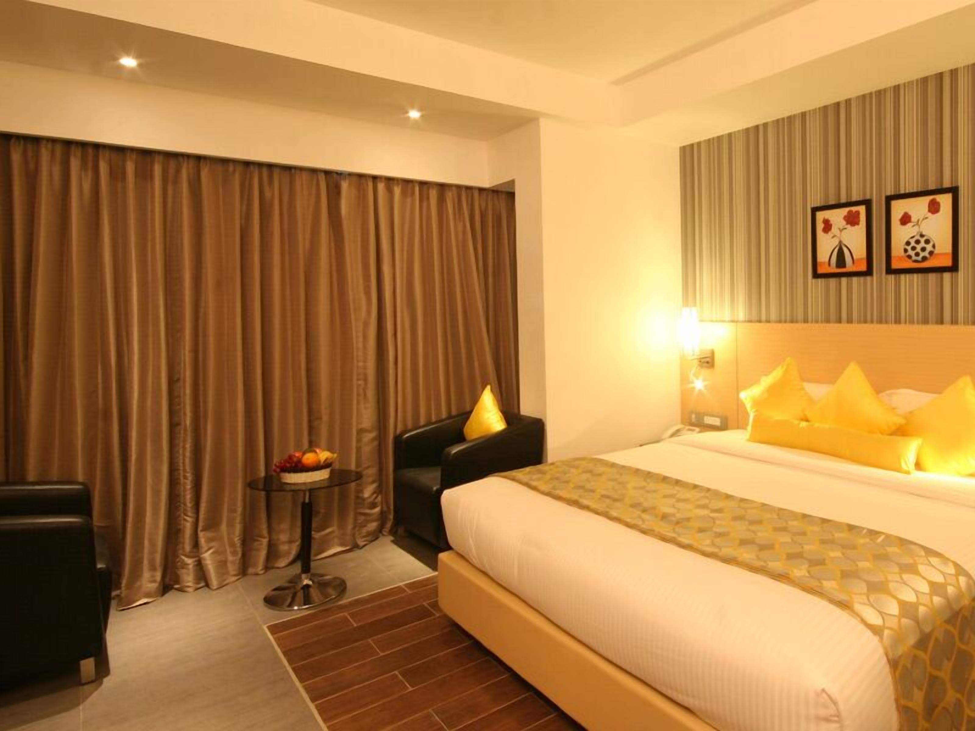 A hotel room with a large bed, a small table with fruit, a chair, and a wall with two framed pictures.