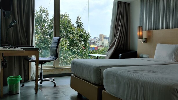 A hotel room with two beds, a desk, a chair, and a view of the city. - Landmark Residency (Mumbai)