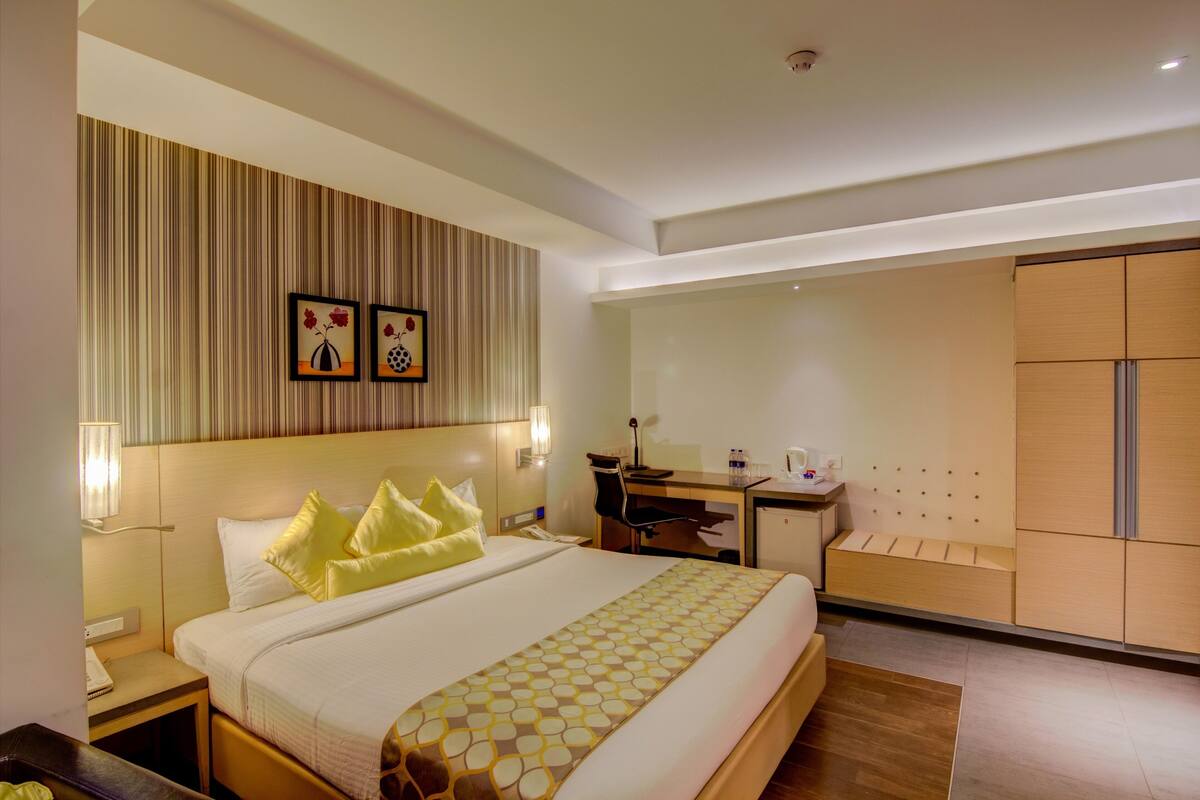 A hotel room with a large bed, a desk, a chair, a wardrobe, and two framed pictures on the wall.