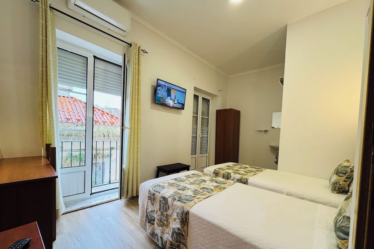 A hotel room with two beds, a TV on the wall, a wardrobe, and a balcony with a view of buildings.
