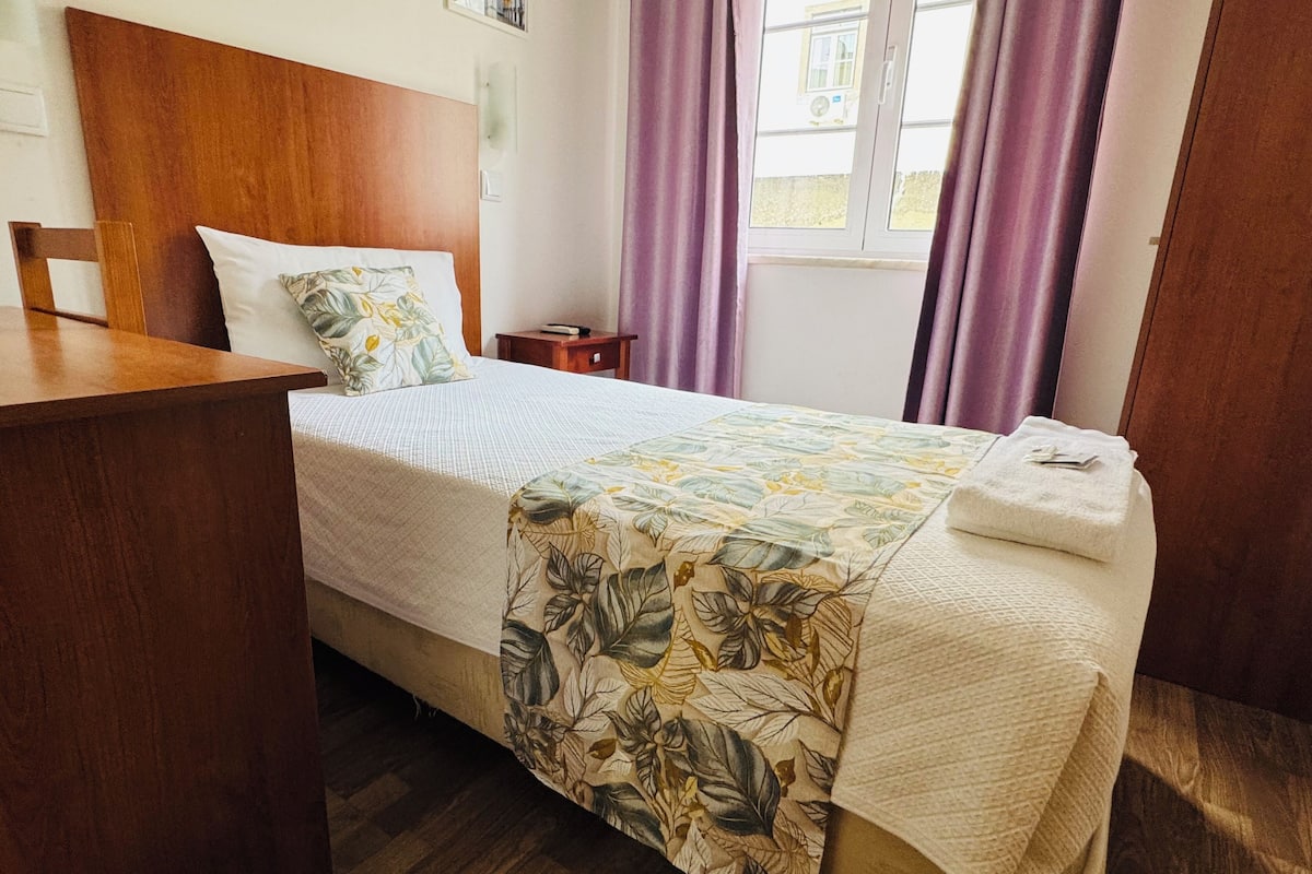 A neatly made bed with a floral bedspread, a wooden headboard, a small bedside table, and a window with purple curtains.