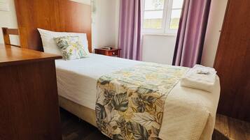 A neatly made bed with a floral bedspread, a wooden headboard, a small bedside table, and a window with purple curtains.