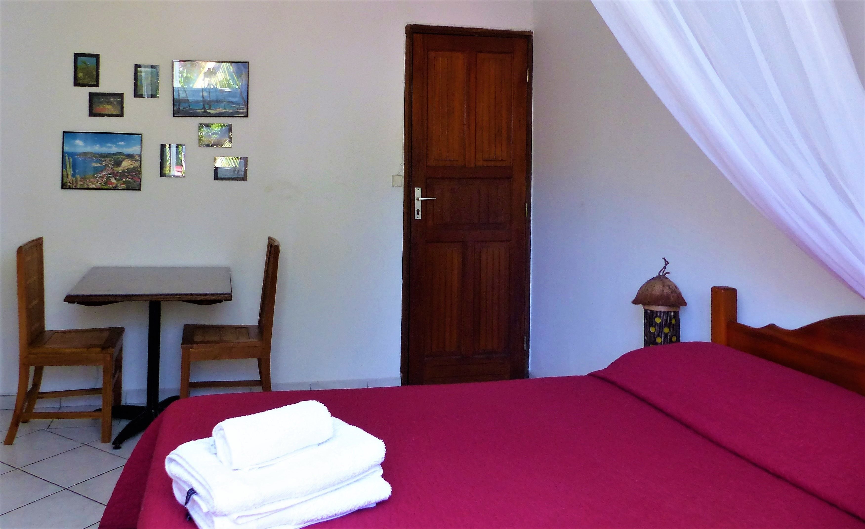 A bedroom with a bed, a wooden door, a table with two chairs, and several framed pictures on the wall.
