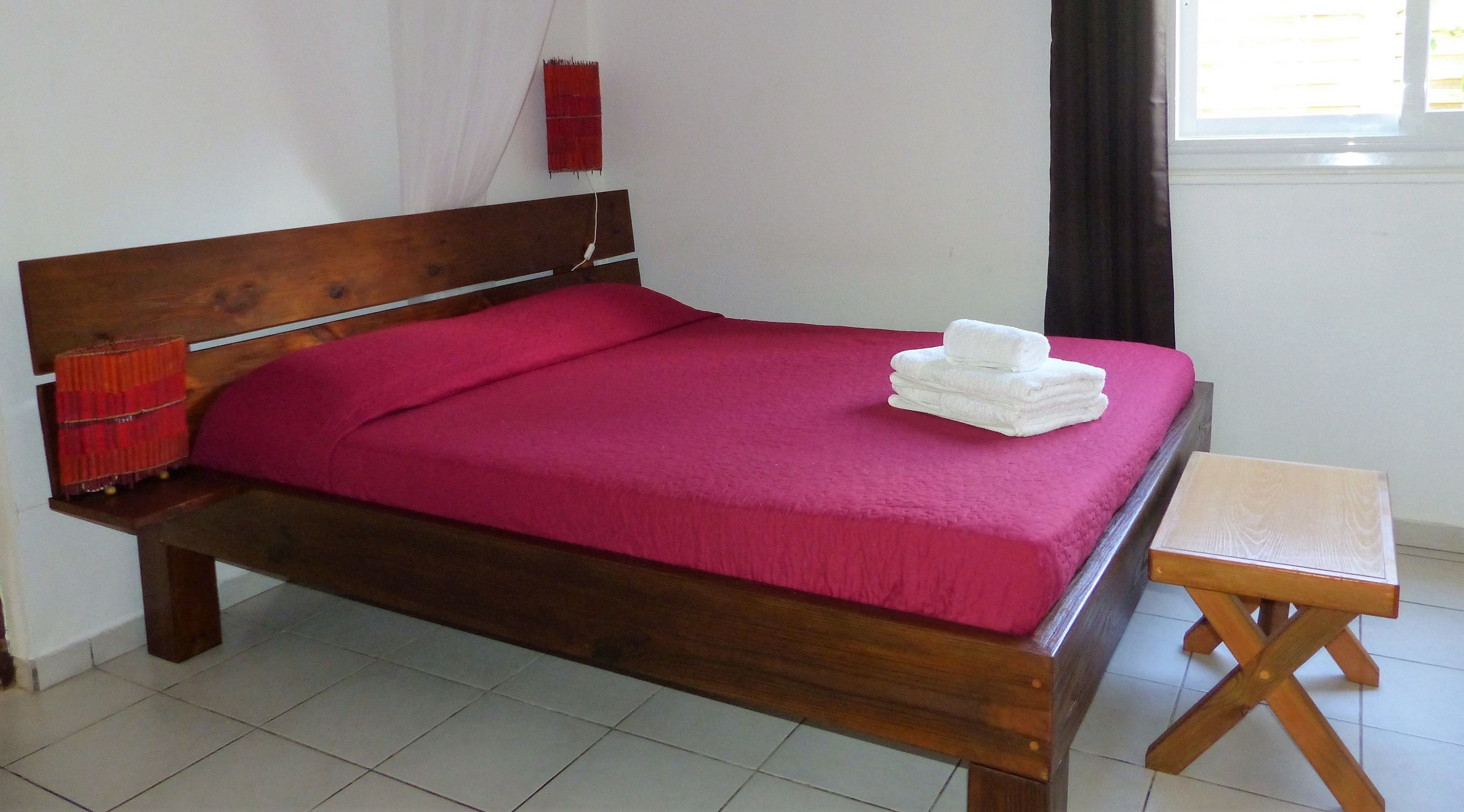 A wooden bed with a red bedspread and white towels, a small wooden stool, and a red decorative item on the nightstand.