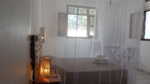 Una habitación con cama, mesita de noche con lámpara, ventana con contraventanas y mosquitera.