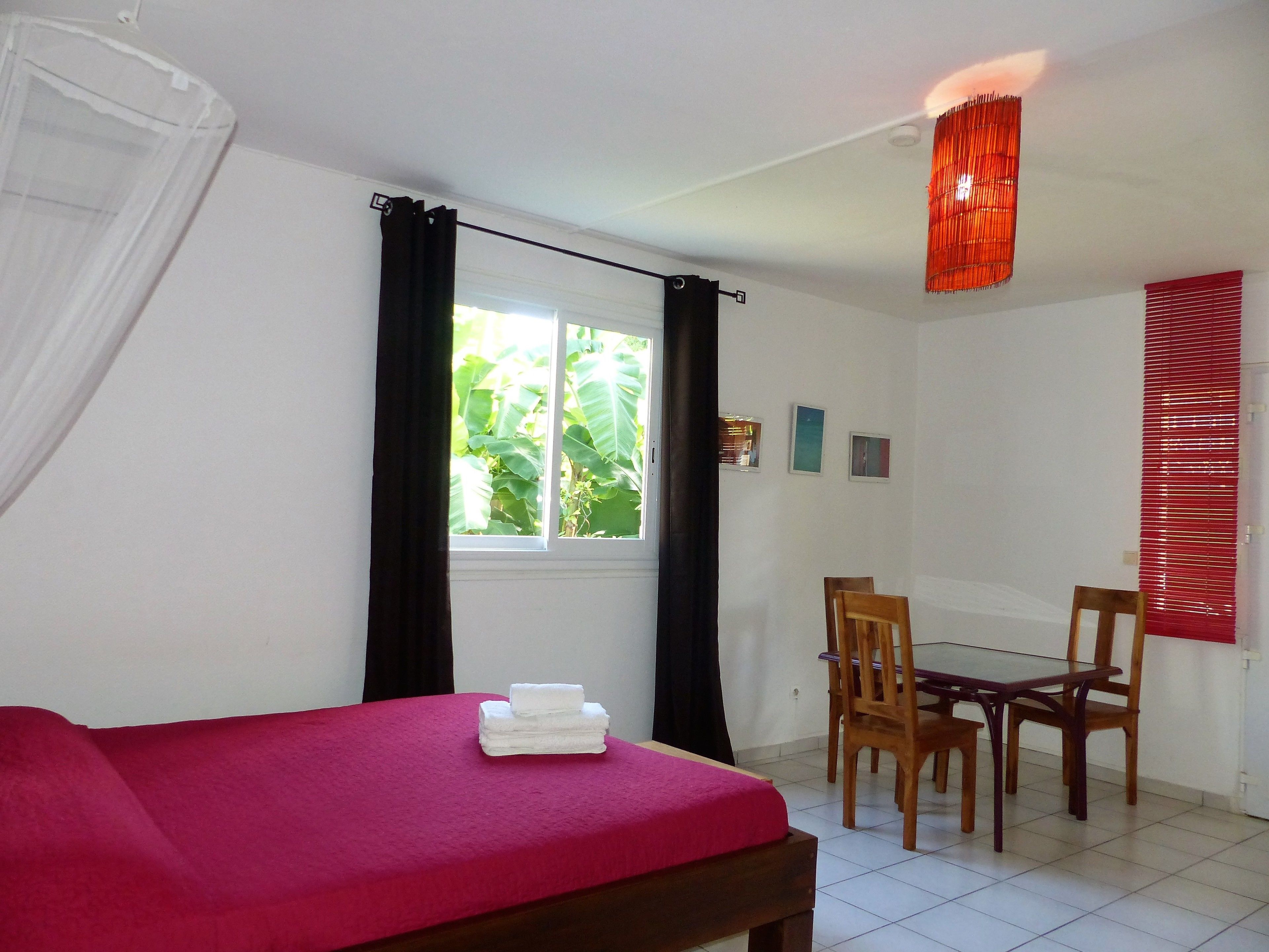 Una habitación con cama, una mesa con sillas, un ventanal con cortinas y una lámpara colgante roja.
