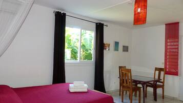 Una habitación con cama, una mesa con sillas, un ventanal con cortinas y una lámpara colgante roja.