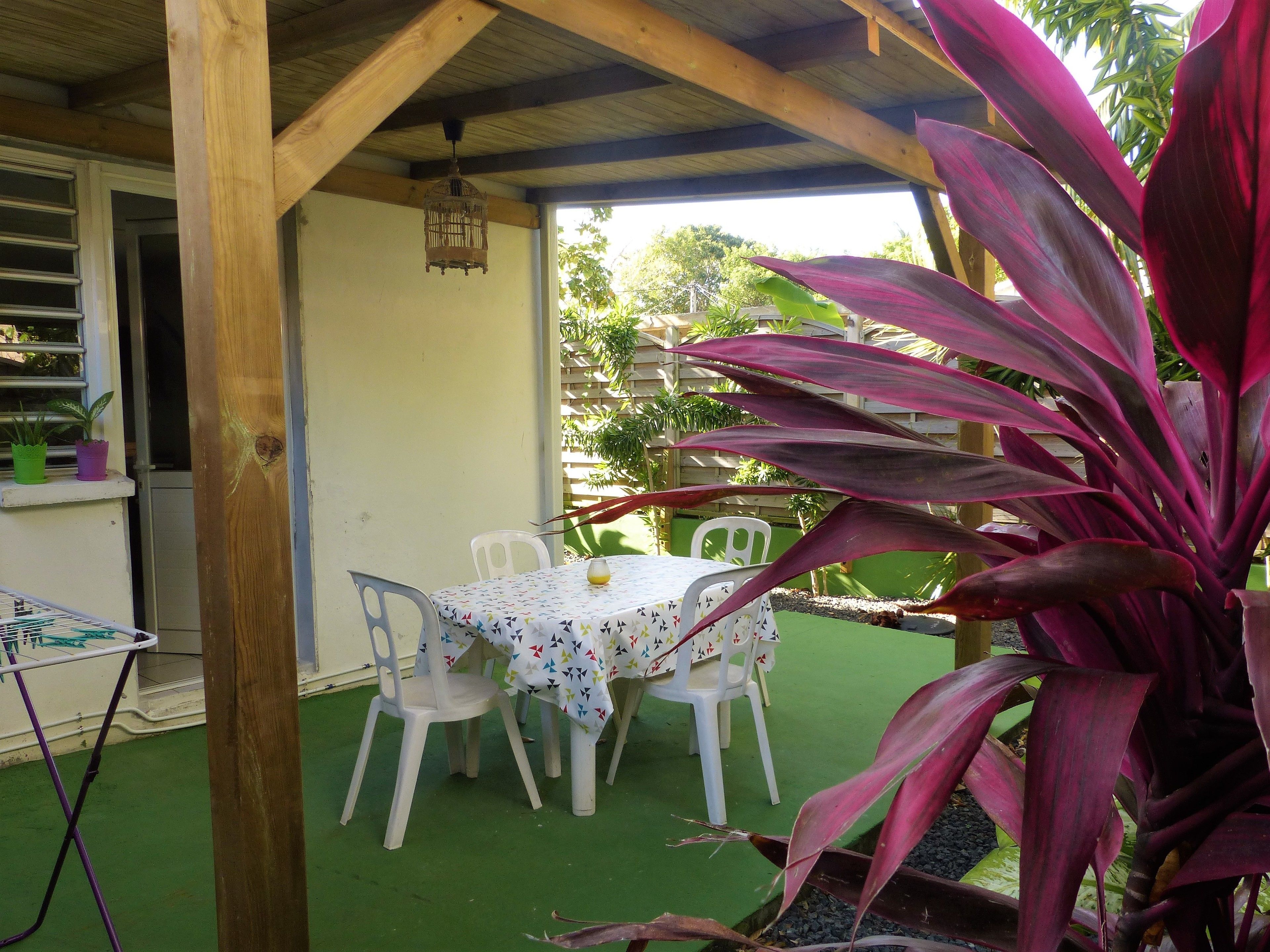 An outdoor patio with a wooden pergola, a table with a patterned tablecloth, and white plastic chairs.