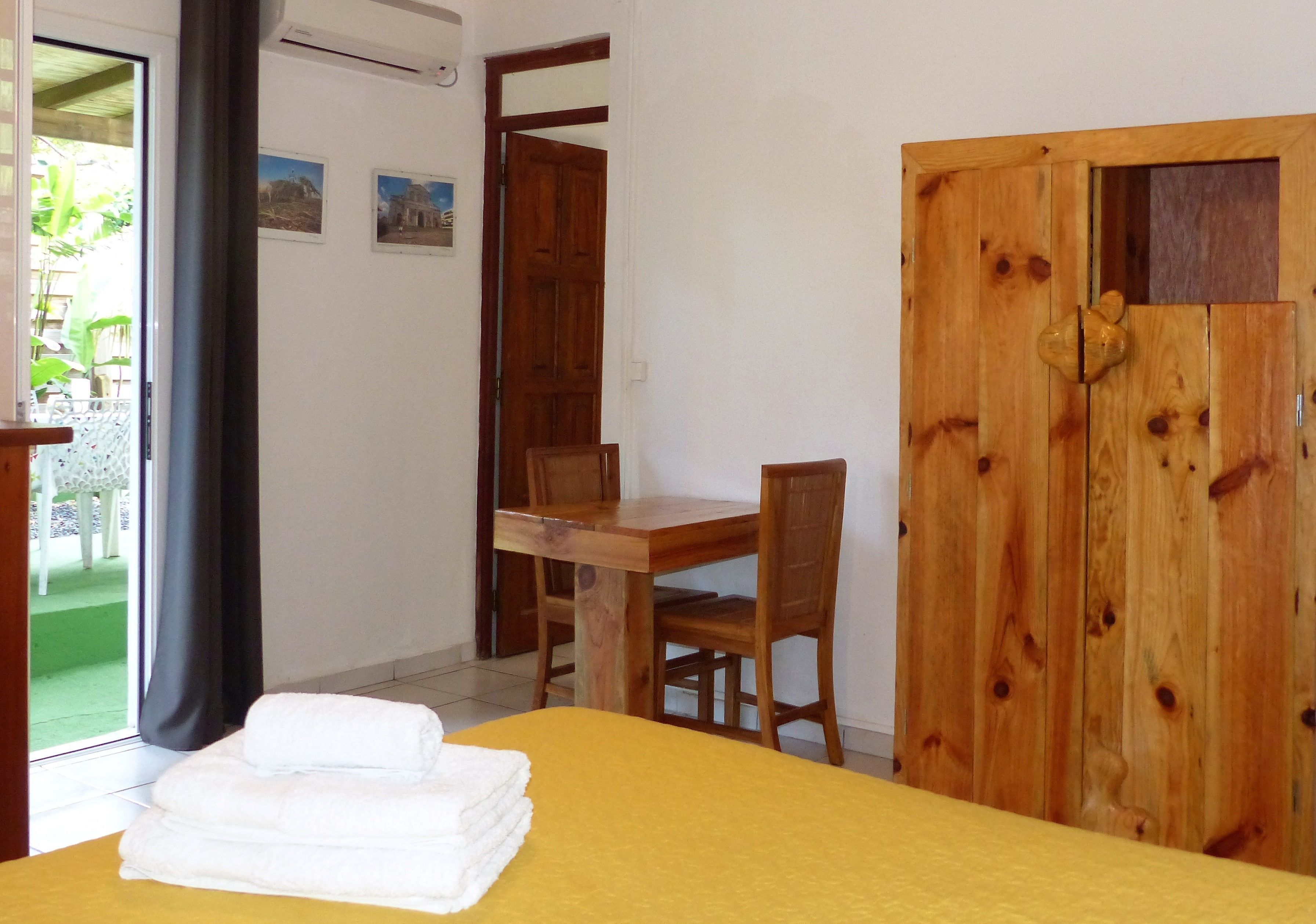 A room with a bed, a wooden door, a dining table with chairs, and a view of the outside.
