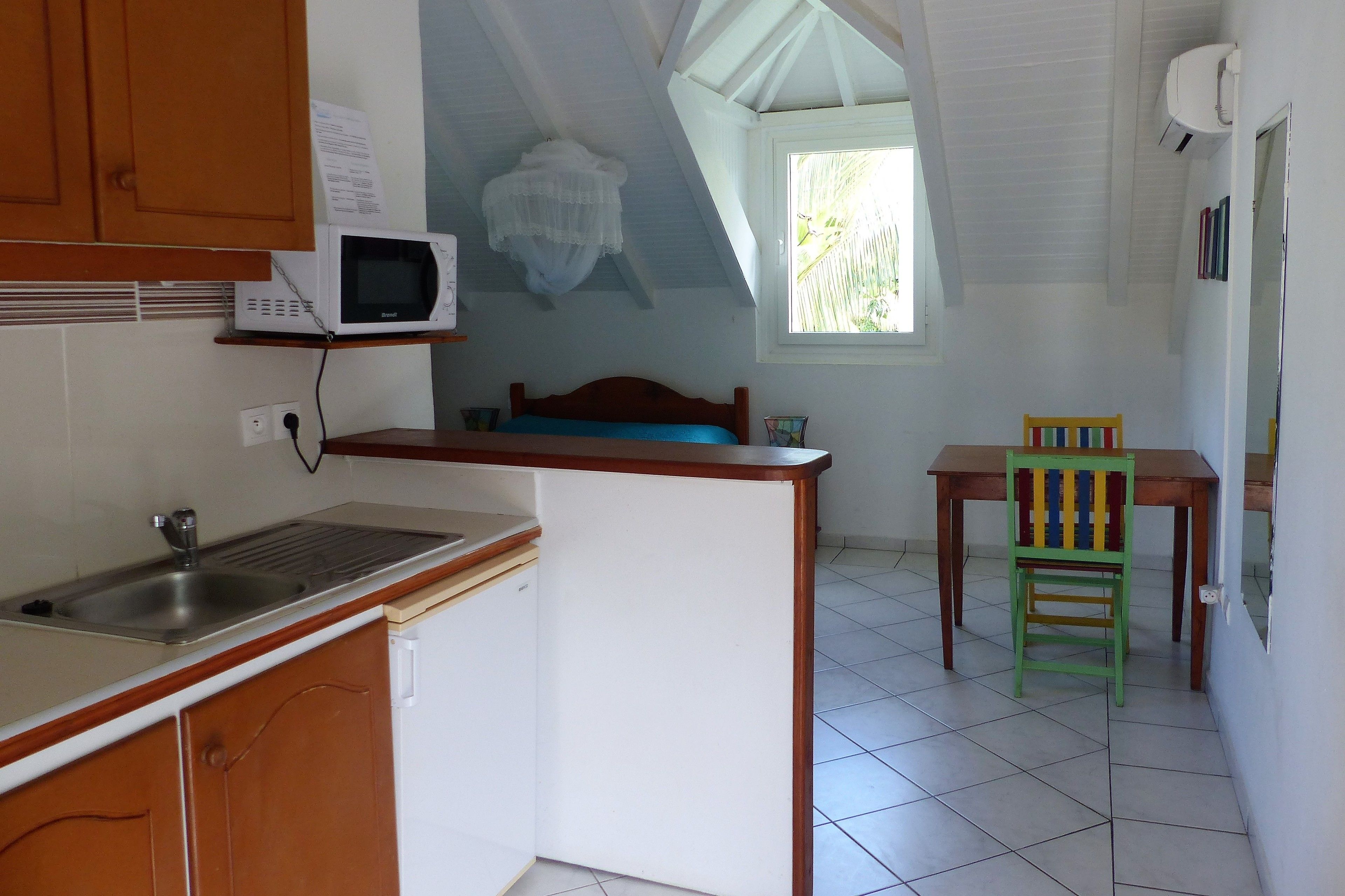 A compact kitchen with a microwave, sink, and wooden cabinets. There is a window providing natural light and a small table with a colorful chair.
