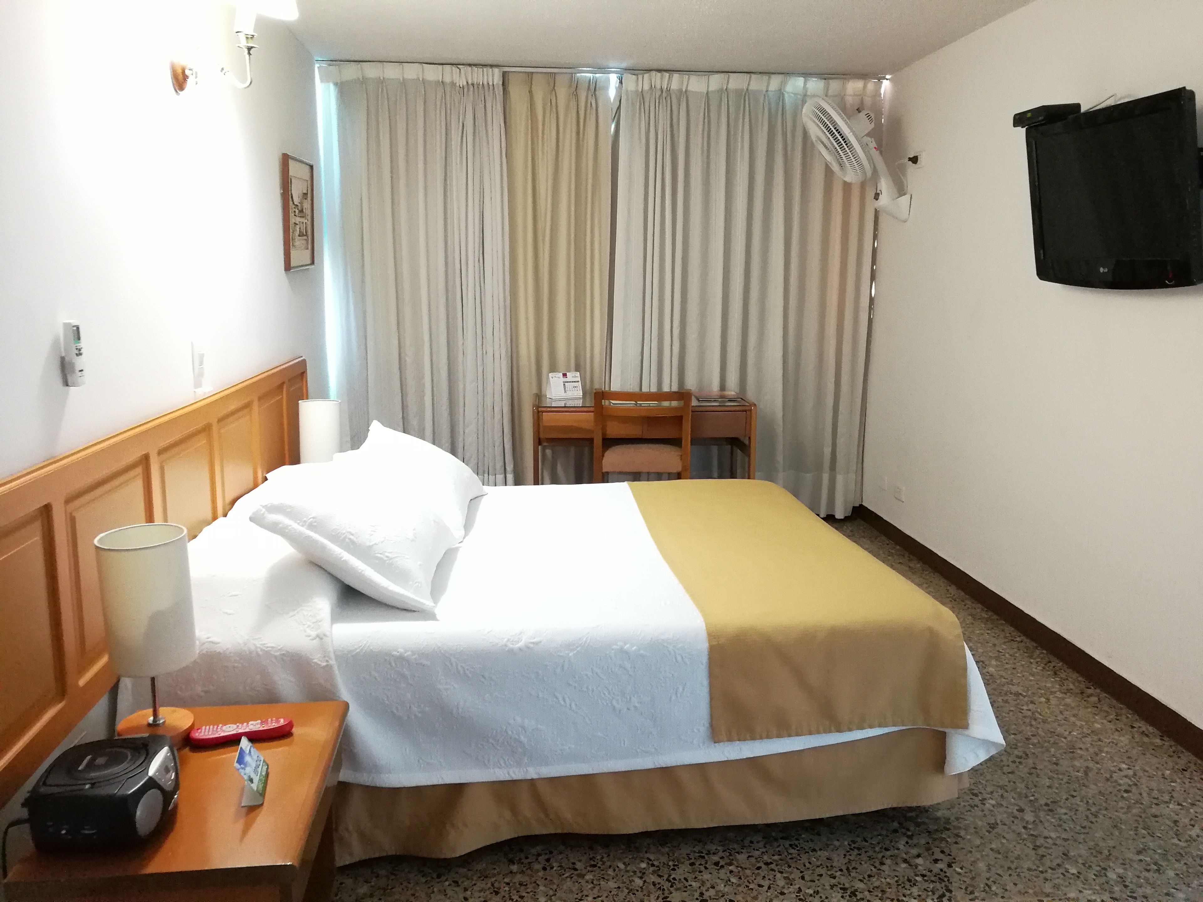 A hotel room with a bed, a desk, a chair, a lamp, a fan, and a wall-mounted TV.