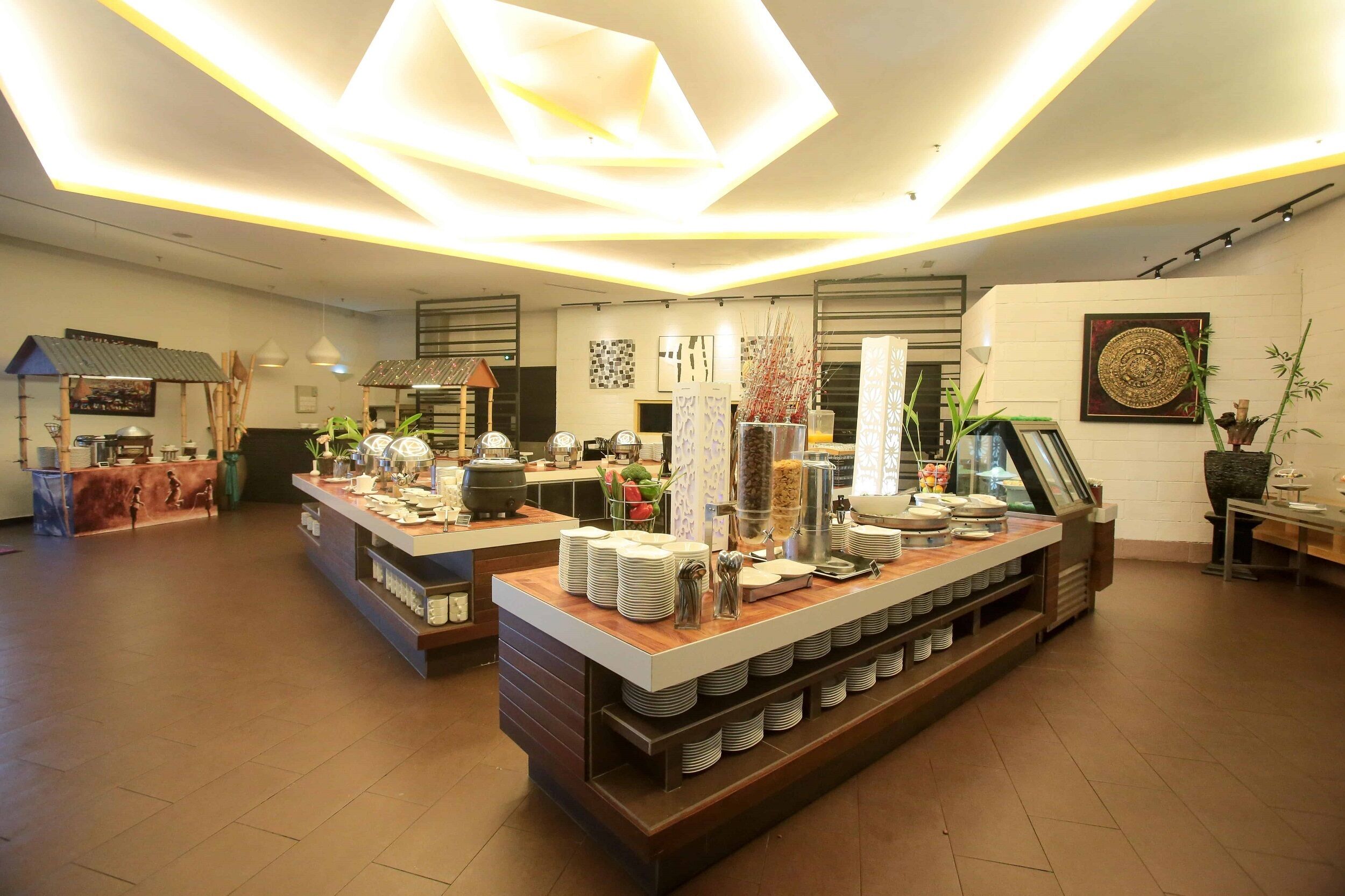 A modern hotel lobby with a central buffet area, a reception desk, and a large decorative wall piece.