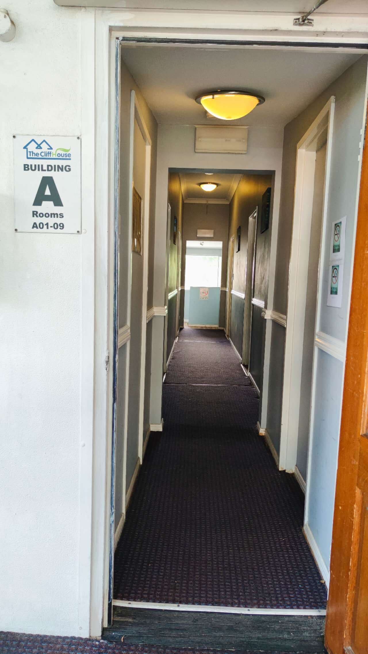 Un couloir étroit, recouvert de moquette, avec des portes de chaque côté, menant à différentes pièces. Une pancarte indique le bâtiment A, pièces A01 à A09.