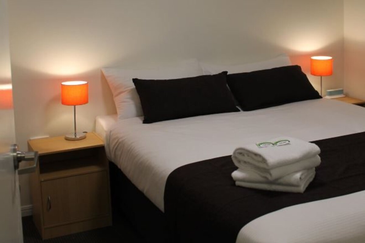 Un lit bien fait, avec du linge de lit blanc, deux oreillers noirs et une serviette blanche pliée posée sur le lit. On y trouve deux lampes de chevet avec des abat-jour orange, ainsi qu’une table de chevet en bois avec un tiroir situé en dessous du lit.